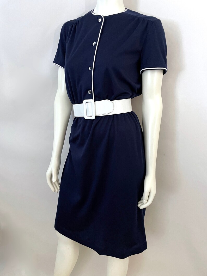 Op de afbeelding: Een marineblauwe jurk met witte bies en bijpassende witte riem. De jurk met korte mouwen heeft een knoopsluiting aan de voorkant en een zoom tot op de knie. De jurk wordt op een mannequin getoond.