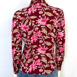 May include: Burgundy button-down shirt with a floral pattern. The shirt features a design of pink flowers and tan leaves. The long-sleeved shirt is paired with blue denim jeans. The shirt is displayed on a mannequin.