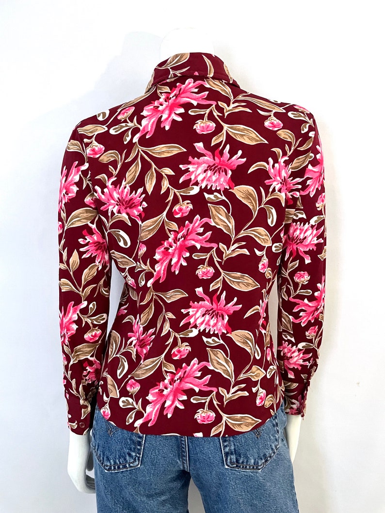 May include: Burgundy button-down shirt with a floral pattern. The shirt features a design of pink flowers and tan leaves. The long-sleeved shirt is paired with blue denim jeans. The shirt is displayed on a mannequin.