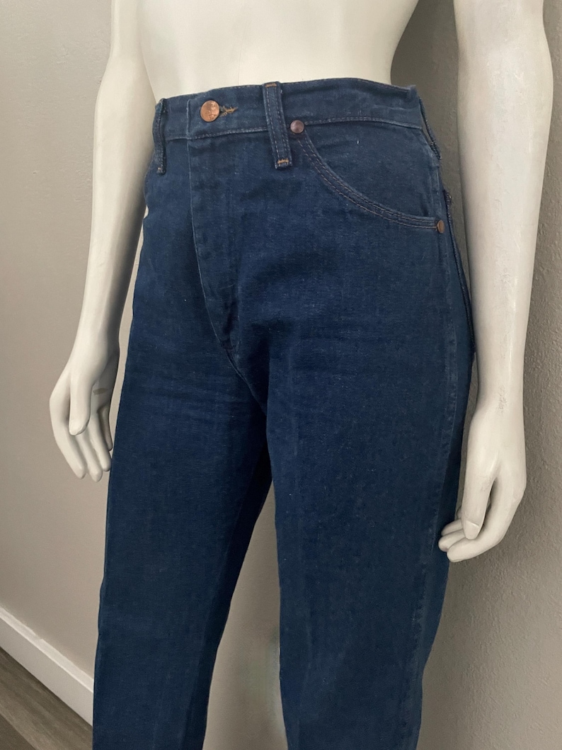 May include: Dark blue denim jeans with a classic five-pocket design. The jeans have copper-colored rivets and a button closure. The fabric appears to be a medium weight denim, suitable for everyday wear. The jeans are displayed on a mannequin.