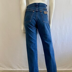 May include: Blue denim jeans with a straight leg fit. The jeans have a classic five-pocket design, a brown leather patch on the back, and a Wrangler label. The jeans are displayed on a mannequin.