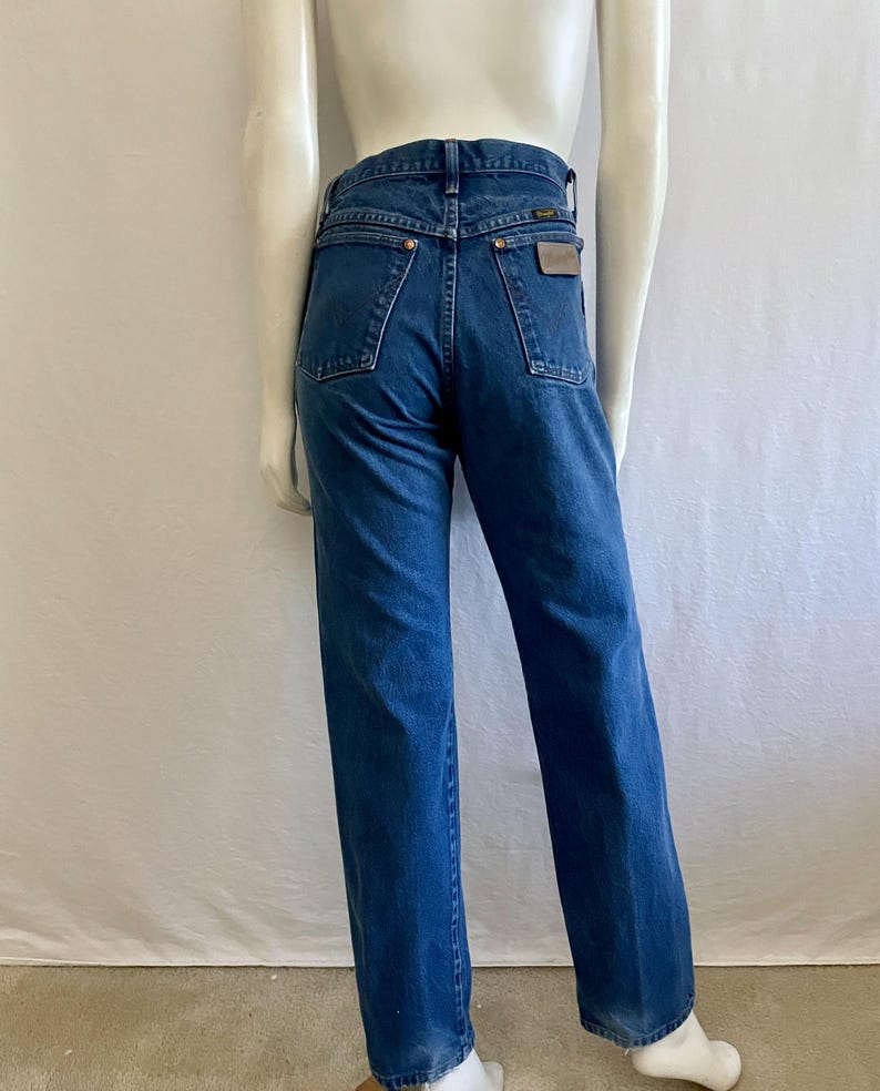 May include: Blue denim jeans with a straight leg fit. The jeans have a classic five-pocket design, a brown leather patch on the back, and a Wrangler label. The jeans are displayed on a mannequin.