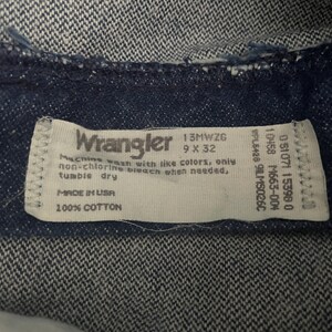 May include: Close-up of a dark blue denim fabric with a white label. The label reads "Wrangler" and includes washing instructions, size 9 x 32, and "Made in USA". The fabric is 100% cotton.