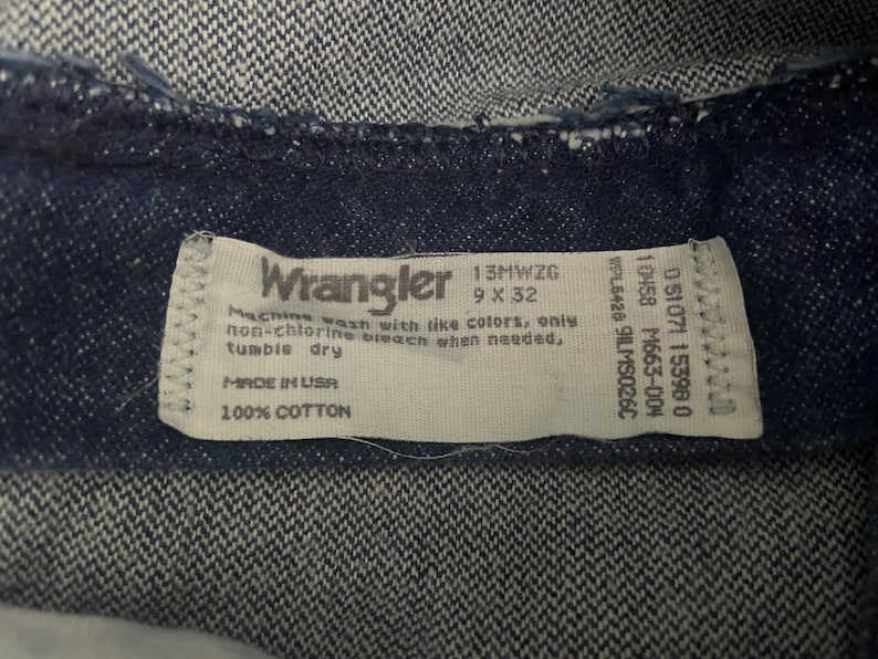 May include: Close-up of a dark blue denim fabric with a white label. The label reads "Wrangler" and includes washing instructions, size 9 x 32, and "Made in USA". The fabric is 100% cotton.