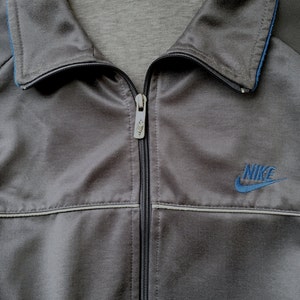 May include: A grey Nike track jacket with a full zip and a blue embroidered Nike logo. The jacket features a collared neckline with blue trim and a light grey lining. The jacket is made of a shiny, smooth material.