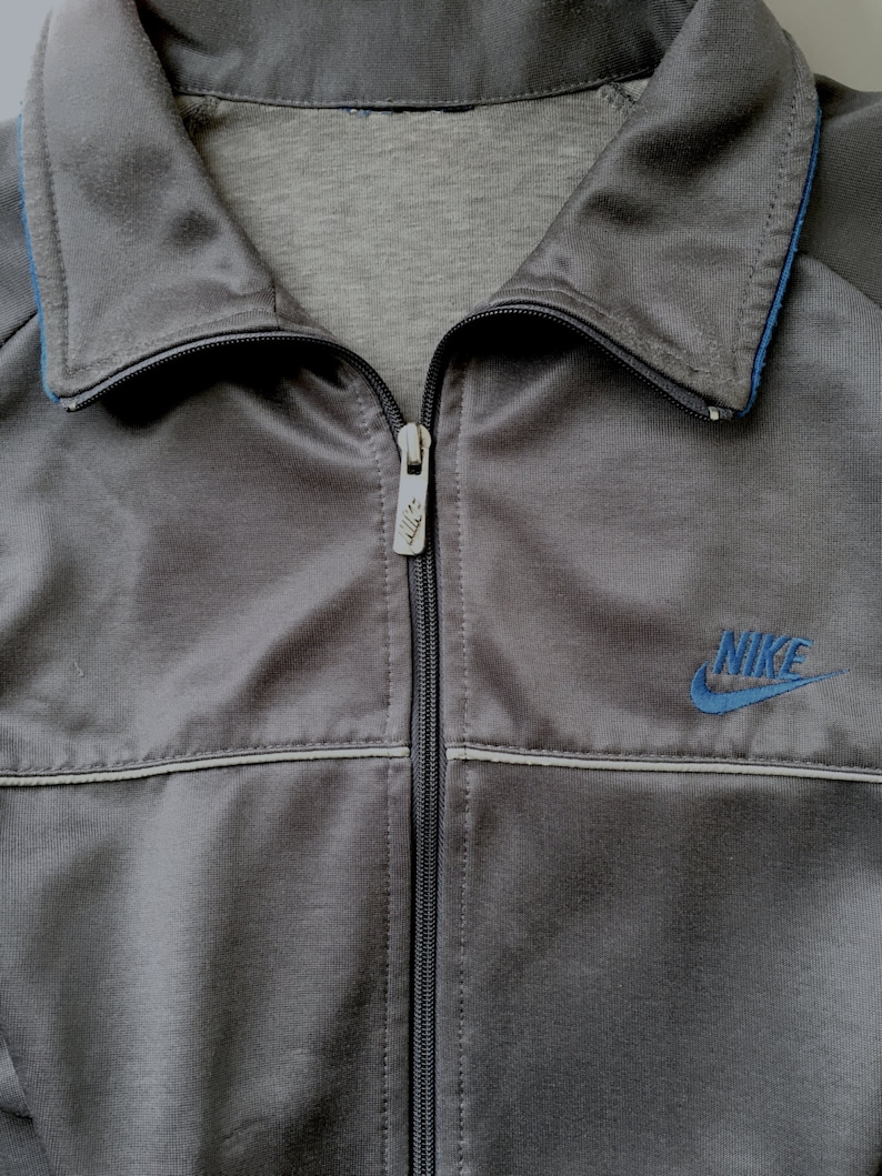 May include: A grey Nike track jacket with a full zip and a blue embroidered Nike logo. The jacket features a collared neckline with blue trim and a light grey lining. The jacket is made of a shiny, smooth material.