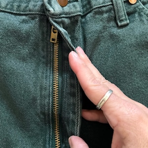 May include: Close-up of dark green denim jeans with a brass zipper and button closure. The jeans have a classic five-pocket design and a worn appearance. The zipper is partially open, revealing the interior of the jeans.