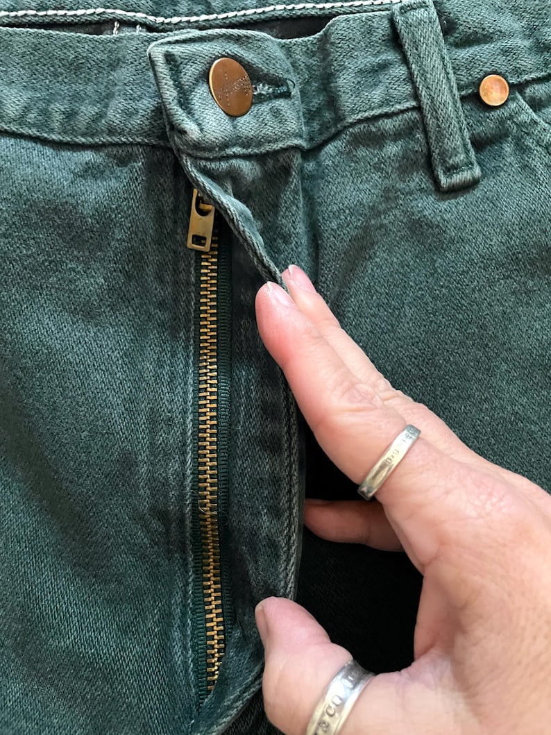 May include: Close-up of dark green denim jeans with a brass zipper and button closure. The jeans have a classic five-pocket design and a worn appearance. The zipper is partially open, revealing the interior of the jeans.
