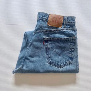 May include: Light blue denim jeans with a classic five-pocket design. The jeans feature a brown leather patch with text and a red Levi's tag on the back pocket. The jeans are folded neatly.