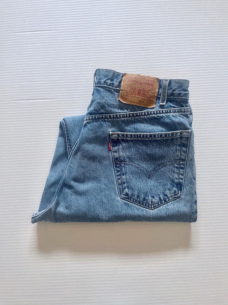 May include: Light blue denim jeans with a classic five-pocket design. The jeans feature a brown leather patch with text and a red Levi's tag on the back pocket. The jeans are folded neatly.
