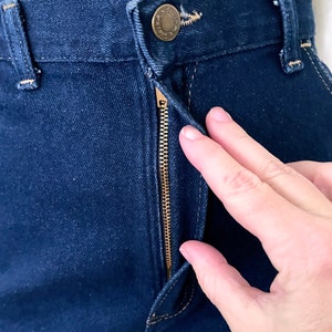 K&ouml;nnte beinhalten: Nahaufnahme einer dunkelblauen Jeans mit Messingrei&szlig;verschluss und Knopfverschluss. Die Jeans hat sichtbare N&auml;hte und ein klassisches F&uuml;nf-Taschen-Design. Der Rei&szlig;verschluss ist teilweise ge&ouml;ffnet, und eine Hand ist in der N&auml;he des Verschlusses zu sehen.