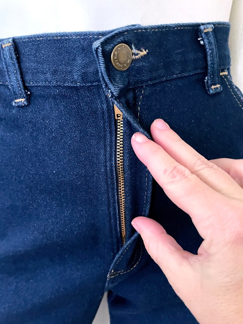 K&ouml;nnte beinhalten: Nahaufnahme einer dunkelblauen Jeans mit Messingrei&szlig;verschluss und Knopfverschluss. Die Jeans hat sichtbare N&auml;hte und ein klassisches F&uuml;nf-Taschen-Design. Der Rei&szlig;verschluss ist teilweise ge&ouml;ffnet, und eine Hand ist in der N&auml;he des Verschlusses zu sehen.