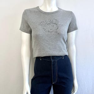 May include: A grey t-shirt with a crew neck and short sleeves. The shirt has a black graphic of a moon with the text "The Star Tree" above it. The shirt is worn with dark blue jeans.
