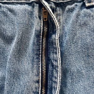 May include: Close-up of light blue denim jeans with a brass zipper. The zipper is partially open, revealing the teeth and the fabric's texture. The jeans show signs of wear, with visible stitching and a button.