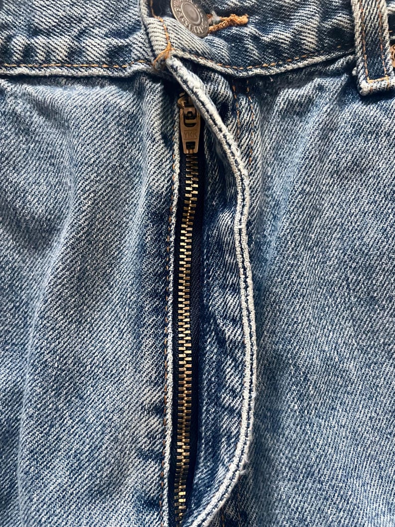 May include: Close-up of light blue denim jeans with a brass zipper. The zipper is partially open, revealing the teeth and the fabric's texture. The jeans show signs of wear, with visible stitching and a button.