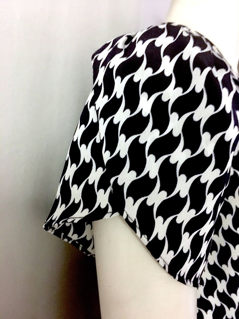 May include: A black and white patterned top with a repeating wave design. The fabric is draped over a mannequin, showcasing the sleeve and shoulder detail. The pattern is a bold, graphic design.