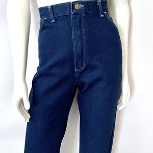 K&ouml;nnte beinhalten: Dunkelblaue Jeans mit hohem Bund und Knopfverschluss. Die Jeans haben ein klassisches F&uuml;nf-Taschen-Design und werden auf einer Schaufensterpuppe pr&auml;sentiert. Die N&auml;hte sind in einer kontrastierenden hellen Farbe.