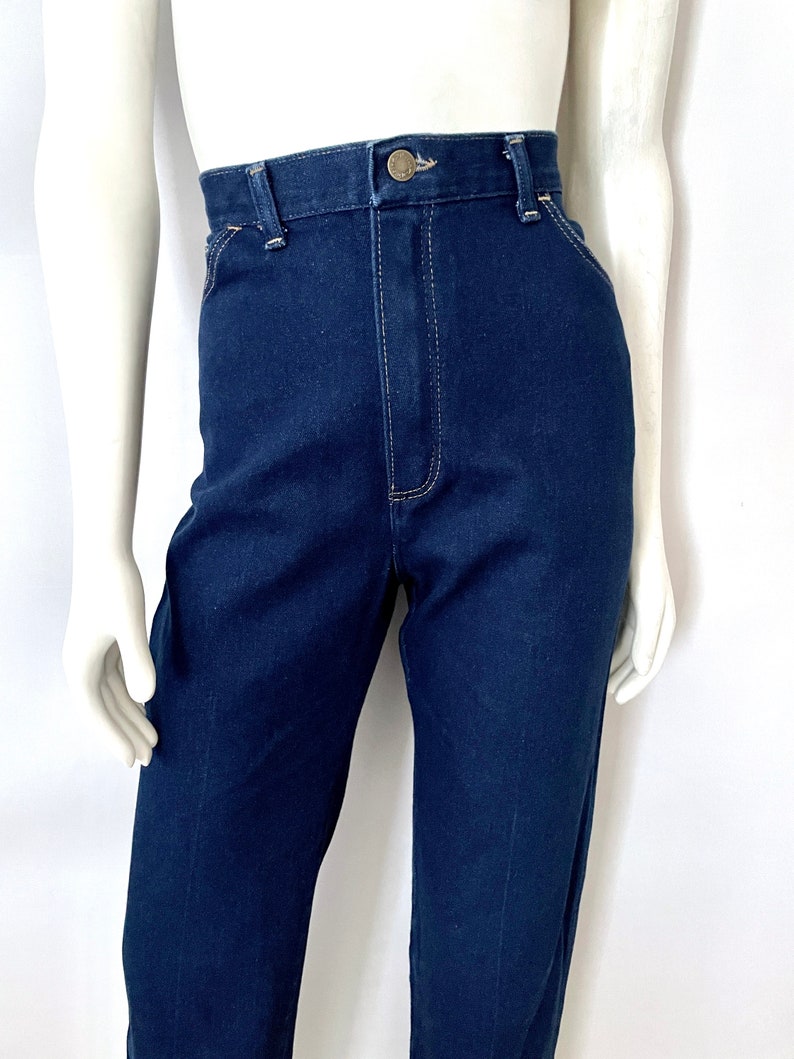 K&ouml;nnte beinhalten: Dunkelblaue Jeans mit hohem Bund und Knopfverschluss. Die Jeans haben ein klassisches F&uuml;nf-Taschen-Design und werden auf einer Schaufensterpuppe pr&auml;sentiert. Die N&auml;hte sind in einer kontrastierenden hellen Farbe.