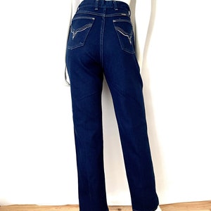 K&ouml;nnte beinhalten: Dunkelblaue Jeans mit hoher Taille und geradem Bein. Die Ges&auml;&szlig;taschen haben ein dekoratives wei&szlig;es Nahtdesign. Die Jeans werden auf einer Schaufensterpuppe vor wei&szlig;em Hintergrund und Holzboden pr&auml;sentiert.