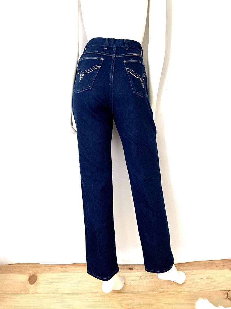 K&ouml;nnte beinhalten: Dunkelblaue Jeans mit hoher Taille und geradem Bein. Die Ges&auml;&szlig;taschen haben ein dekoratives wei&szlig;es Nahtdesign. Die Jeans werden auf einer Schaufensterpuppe vor wei&szlig;em Hintergrund und Holzboden pr&auml;sentiert.