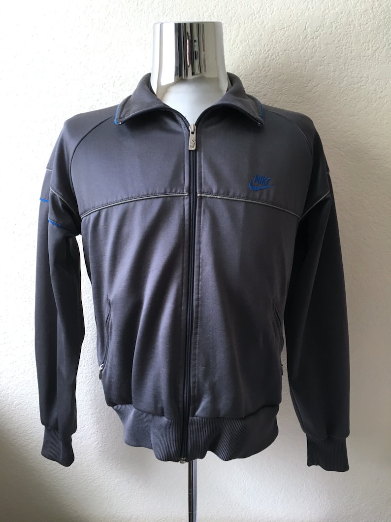 May include: Dark grey track jacket with a full zip. It has a blue Nike logo and blue detailing on the collar and sleeves. The jacket has ribbed cuffs and hem.