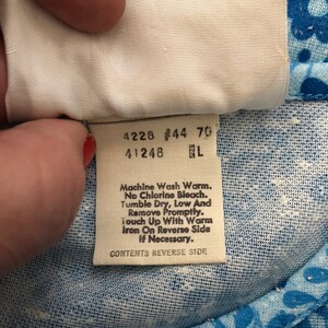 May include: Close-up of a white tag with black text, held against a blue floral fabric. The tag includes washing instructions: Machine Wash Warm, No Chlorine Bleach, Tumble Dry Low, and Iron on Reverse Side if necessary. The tag also has the text "CONTENTS REVERSE SIDE."