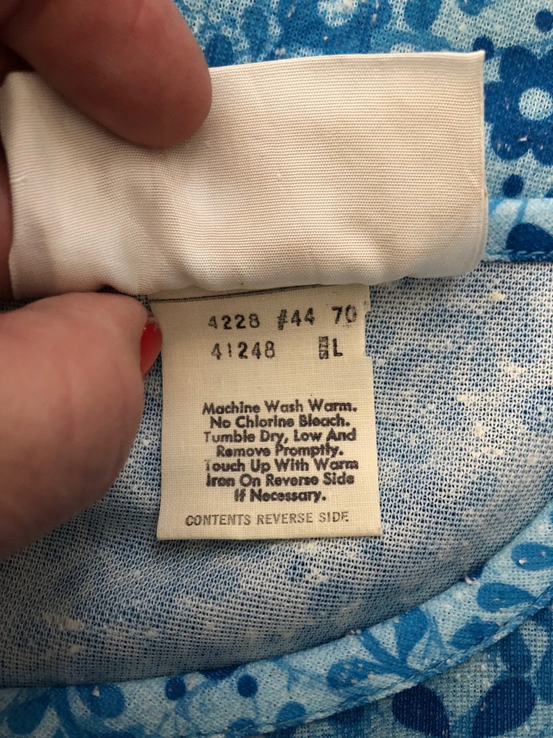 May include: Close-up of a white tag with black text, held against a blue floral fabric. The tag includes washing instructions: Machine Wash Warm, No Chlorine Bleach, Tumble Dry Low, and Iron on Reverse Side if necessary. The tag also has the text "CONTENTS REVERSE SIDE."