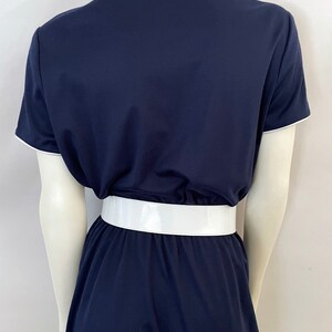 Op de afbeelding: Marineblauwe jurk met korte mouwen en witte bies. De jurk heeft een brede, glanzende witte riem in de taille. De jurk wordt op een mannequin gepresenteerd.