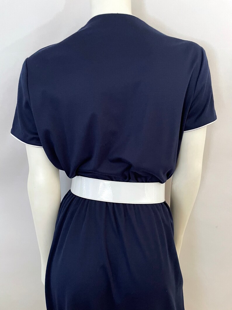 Op de afbeelding: Marineblauwe jurk met korte mouwen en witte bies. De jurk heeft een brede, glanzende witte riem in de taille. De jurk wordt op een mannequin gepresenteerd.