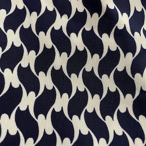 May include: Close-up of a fabric with a repeating pattern. The design features a series of interconnected, stylized shapes in a dark navy blue color against a cream-colored background. The pattern creates a flowing, wave-like effect.