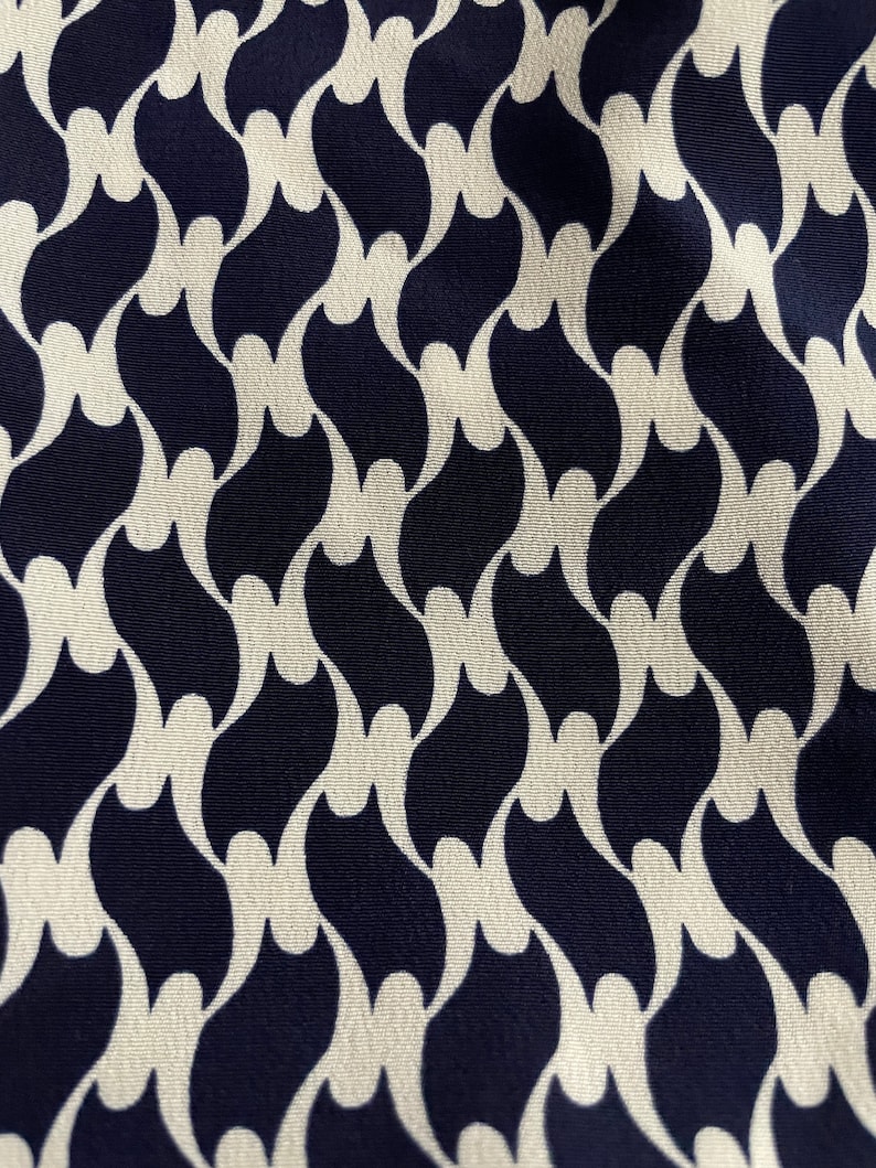 May include: Close-up of a fabric with a repeating pattern. The design features a series of interconnected, stylized shapes in a dark navy blue color against a cream-colored background. The pattern creates a flowing, wave-like effect.