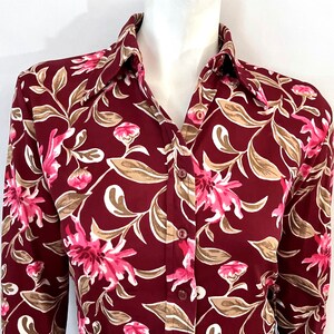 May include: A burgundy button-down shirt with a floral pattern. The design features pink flowers, beige leaves, and white accents. The shirt has a classic collar and long sleeves, suitable for various occasions.