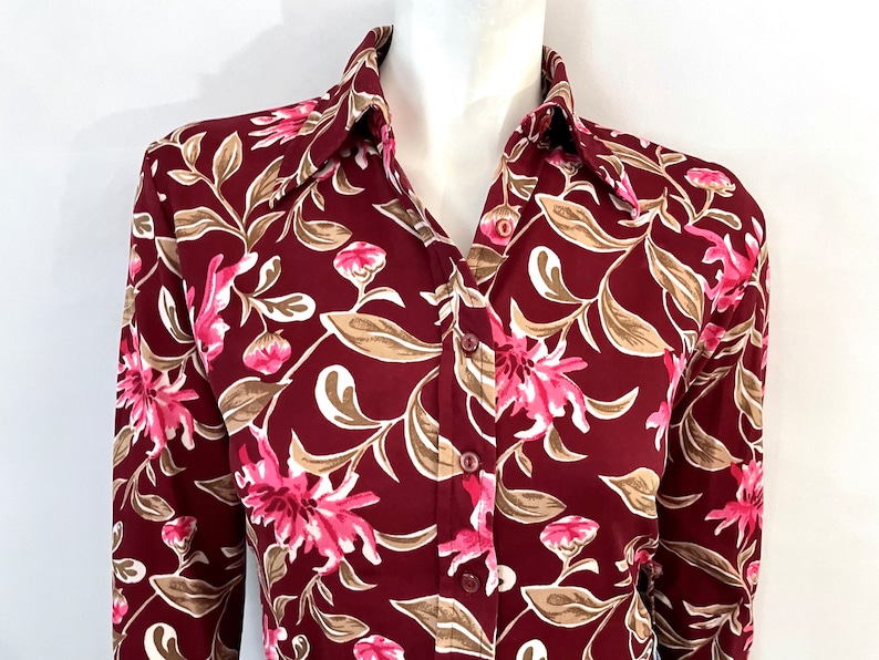 May include: A burgundy button-down shirt with a floral pattern. The design features pink flowers, beige leaves, and white accents. The shirt has a classic collar and long sleeves, suitable for various occasions.