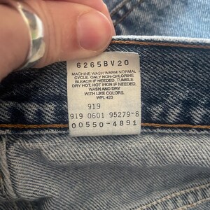 May include: Close-up of a faded blue denim garment. A white tag displays care instructions and product codes: 6265BV20, 919, 919 0601 95279-8, and 00550-4891. The tag also includes washing instructions.