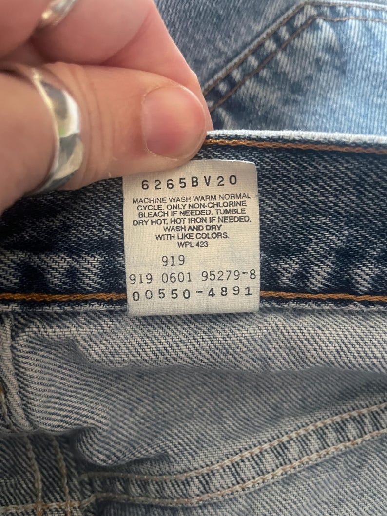 May include: Close-up of a faded blue denim garment. A white tag displays care instructions and product codes: 6265BV20, 919, 919 0601 95279-8, and 00550-4891. The tag also includes washing instructions.