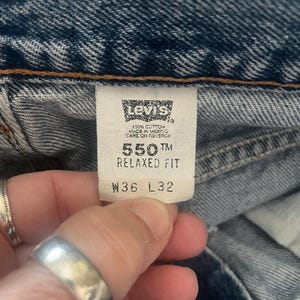 May include: Close-up of a Levi's tag on blue denim jeans. The tag reads "550 Relaxed Fit" and "W36 L32". The jeans are made of 100% cotton and made in Mexico. The tag is held by a person's hand.