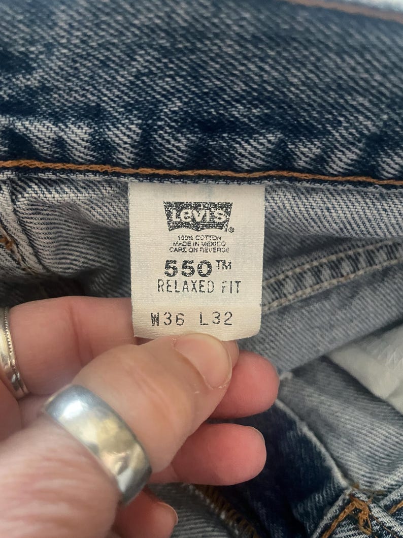May include: Close-up of a Levi's tag on blue denim jeans. The tag reads "550 Relaxed Fit" and "W36 L32". The jeans are made of 100% cotton and made in Mexico. The tag is held by a person's hand.