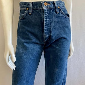 May include: Dark blue denim jeans with a classic five-pocket design. The jeans have a button and zipper closure, belt loops, and copper-colored rivets. The fabric shows subtle fading and wear, giving them a vintage look. The jeans are displayed on a mannequin.