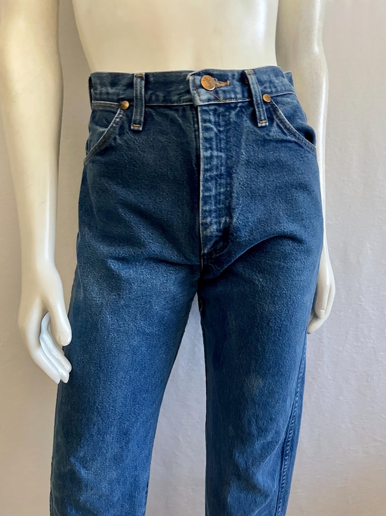 May include: Dark blue denim jeans with a classic five-pocket design. The jeans have a button and zipper closure, belt loops, and copper-colored rivets. The fabric shows subtle fading and wear, giving them a vintage look. The jeans are displayed on a mannequin.