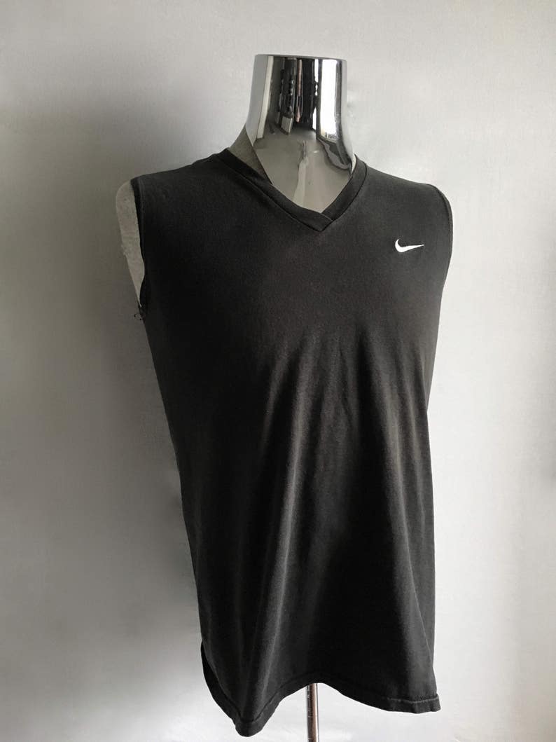 Puede incluir: Camiseta sin mangas gris oscuro con cuello en V y logotipo blanco de Nike en el pecho. La camiseta se muestra en un maniqu&iacute;. La tela parece suave y ligeramente desgastada, lo que sugiere un estilo informal. El aspecto general es sencillo y deportivo.
