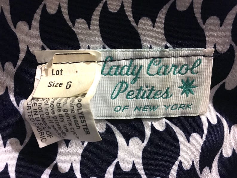 May include: Close-up of a vintage clothing label from "Lady Carol Petites of New York." The label is white with teal lettering and a starburst design. The fabric has a navy and white abstract pattern. A size 6 tag is attached.
