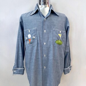 Può includere: Camicia azzurra con maniche lunghe, rifiniture bianche su colletto, polsini e bordi delle tasche. La camicia ha due tasche frontali con disegni ricamati a tema golf. I polsini hanno un bordo smerlato.