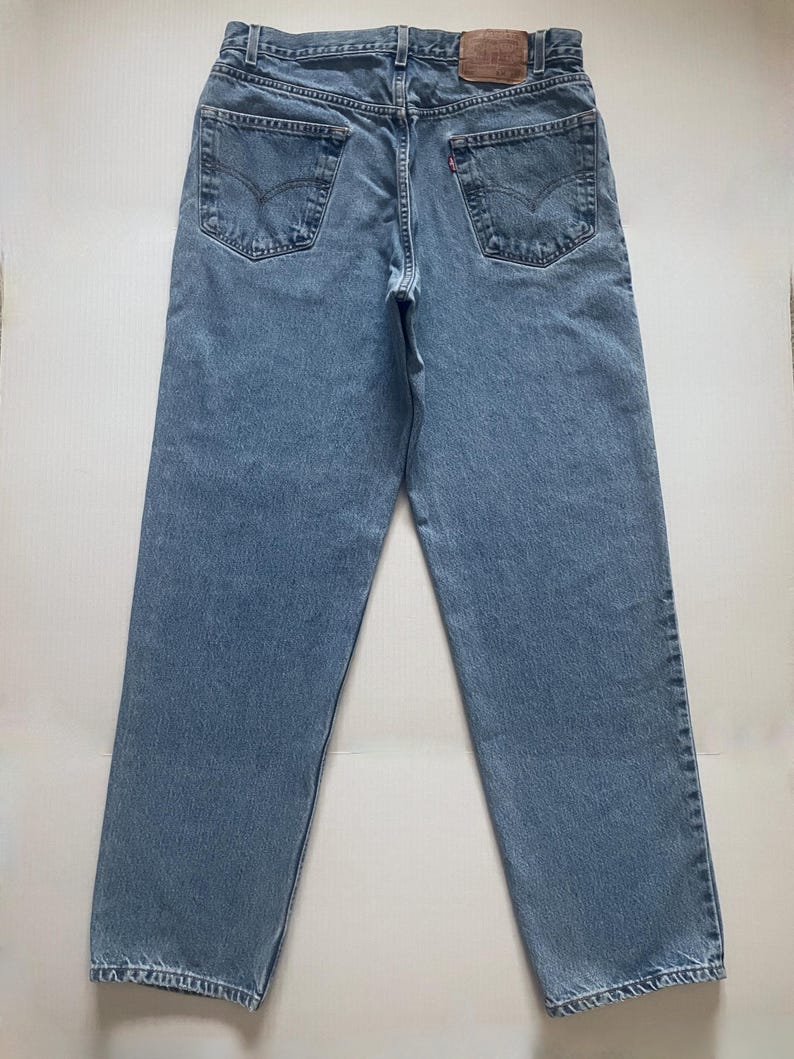 May include: Light blue denim jeans with a classic five-pocket design. The jeans feature a straight leg cut and a leather patch on the back waistband. The back pockets have a signature stitch detail.