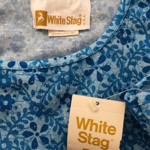 May include: Close-up of a blue floral patterned top with a White Stag tag. The top features a round neckline and a repeating pattern of blue flowers and leaves. The tag displays the White Stag logo and brand name.