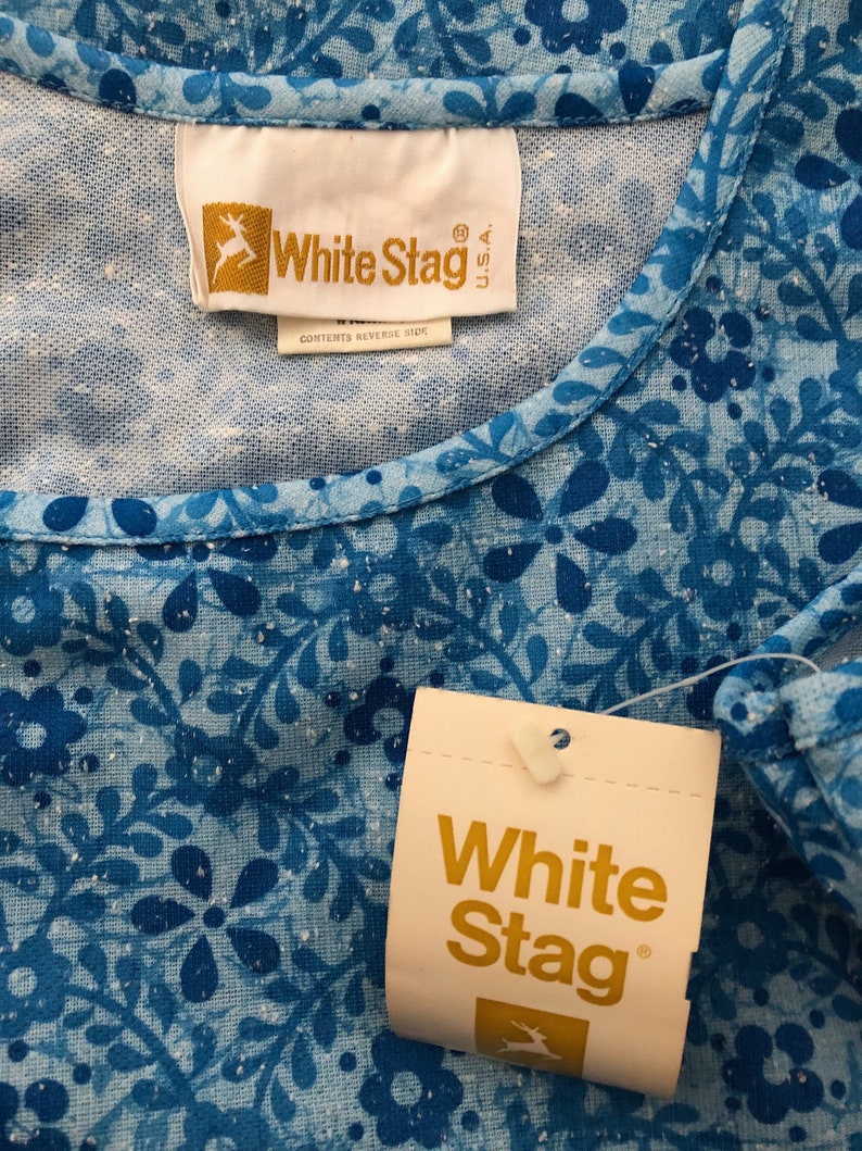 May include: Close-up of a blue floral patterned top with a White Stag tag. The top features a round neckline and a repeating pattern of blue flowers and leaves. The tag displays the White Stag logo and brand name.
