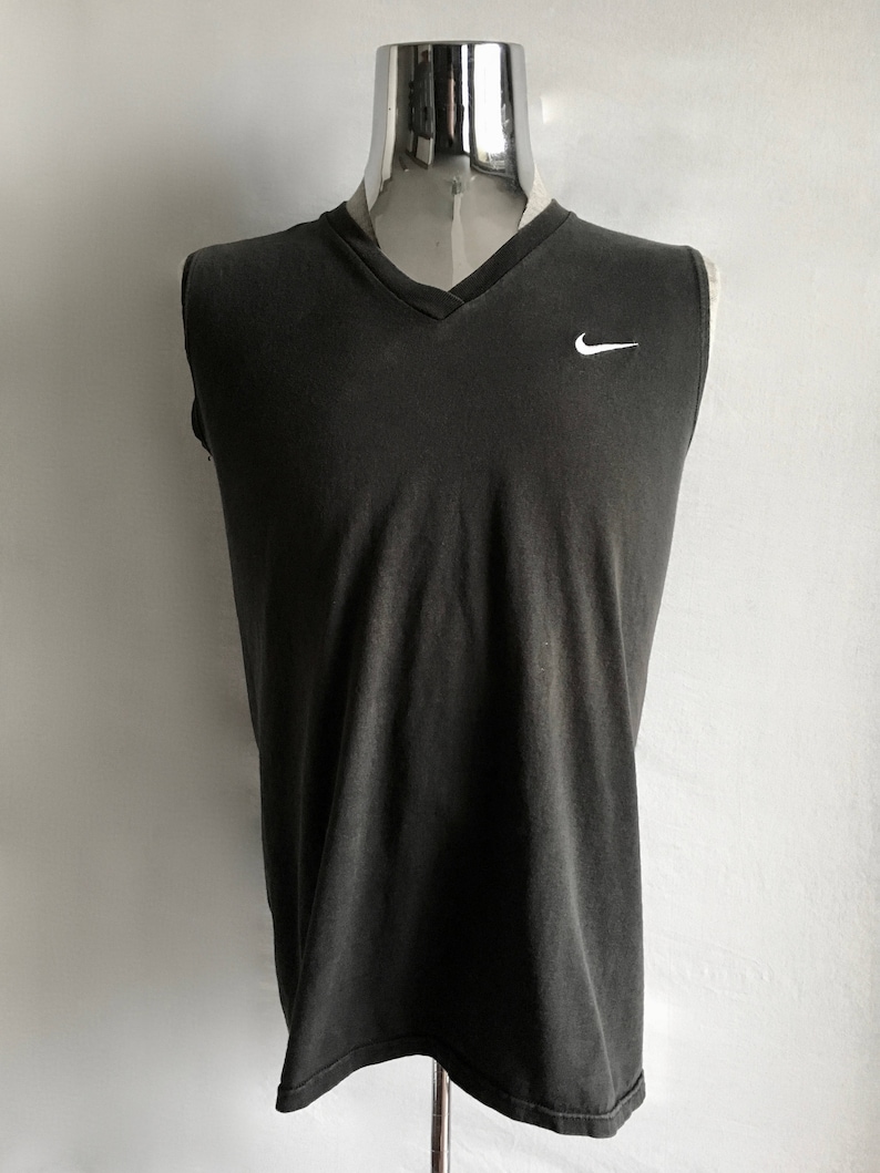 Puede incluir: Camiseta sin mangas gris oscuro con cuello en V. Tiene un peque&ntilde;o logotipo blanco de Nike en el pecho. La prenda se exhibe en un maniqu&iacute;. La tela parece ligeramente descolorida.