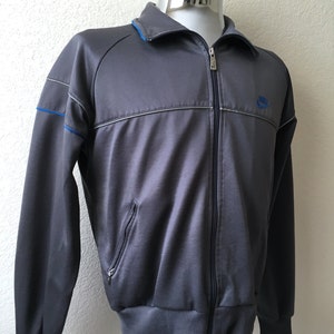 May include: A grey zip-up track jacket with blue collar and accents. The jacket features a small blue Nike logo on the chest and a ribbed hem and cuffs. The jacket has a full zip closure and two front pockets.