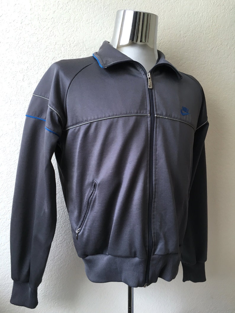 May include: A grey zip-up track jacket with blue collar and accents. The jacket features a small blue Nike logo on the chest and a ribbed hem and cuffs. The jacket has a full zip closure and two front pockets.