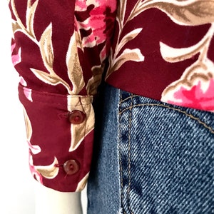 May include: Close-up of a burgundy floral print blouse sleeve with two brown buttons. The floral pattern includes pink and beige flowers and leaves. The sleeve is paired with blue denim jeans.