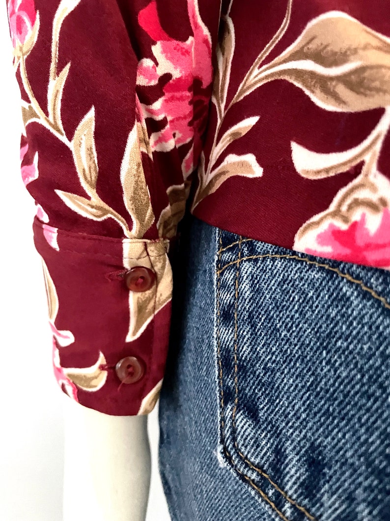 May include: Close-up of a burgundy floral print blouse sleeve with two brown buttons. The floral pattern includes pink and beige flowers and leaves. The sleeve is paired with blue denim jeans.